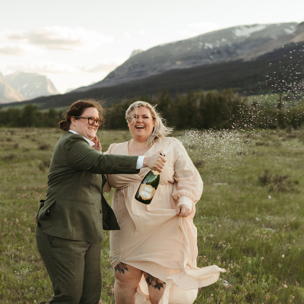 A couple popping a bottle of champagne in a field for their elopement in Glacier National Park.