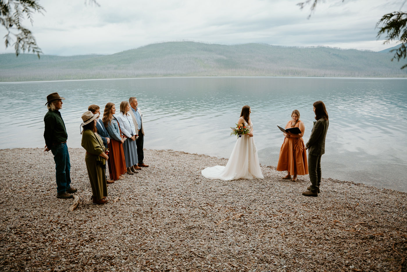 A couple, an officiant, and their guests on the shore of Lake McDonald for their wedding ceremony. Gray clouds cover the sky and the water is slightly rippling in the background.